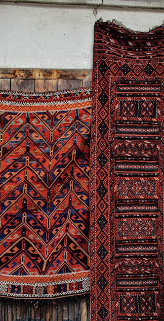 A close-up of an ornate antique rug with intricate floral and geometric patterns in shades of orange, cream, and blue, laid on a wooden floor. Sunlight streams into the room, casting shadows of two bl
