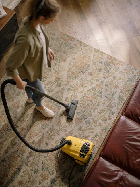 A woman with curly hair, dressed in a green jacket and blue jeans, is performing surface cleaning using a vacuum cleaner on a traditional patterned area rug in a living room. The room features hardwoo